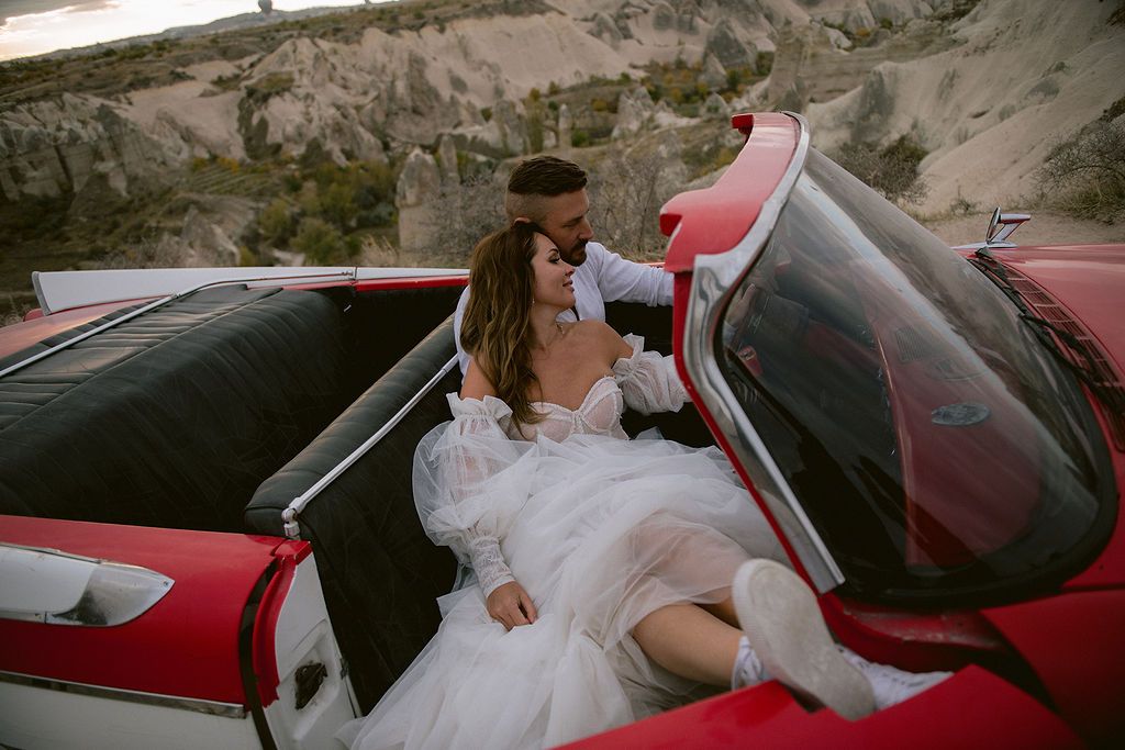A bride and groom share a romantic moment in a vintage red convertible car against a scenic rocky landscape.