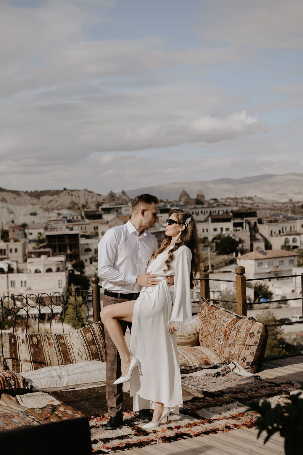 Couple in white attire embracing on a rooftop terrace with scenic Cappadocia landscape and cave dwellings in background.