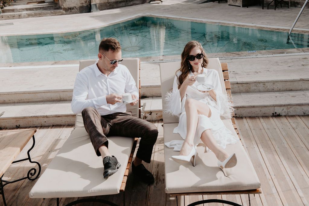 Couple relaxing on poolside loungers, man in white shirt and woman in white dress enjoying drinks on sunny terrace.