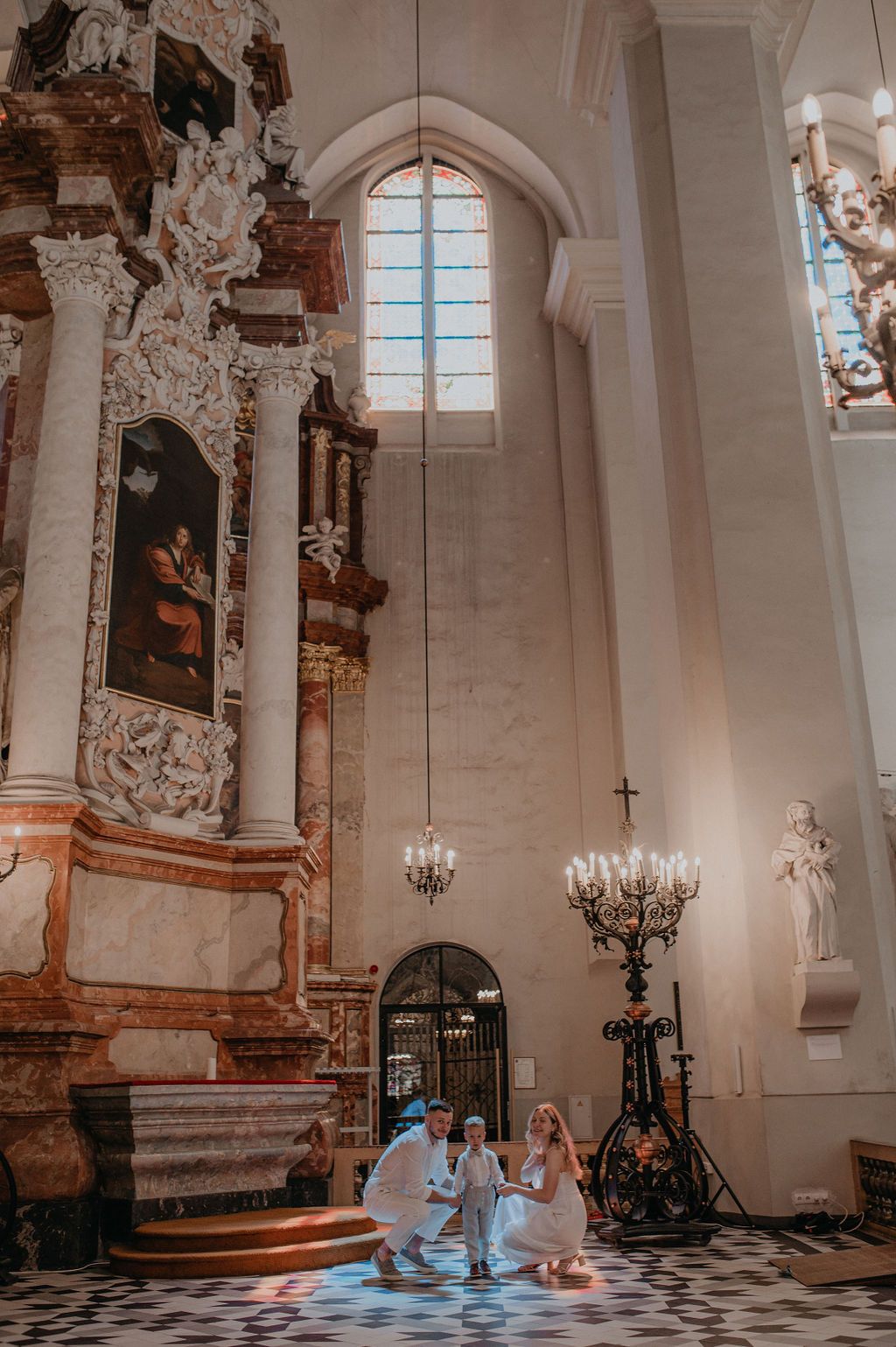 Couple in white attire posing with child inside ornate baroque church with high ceilings and chandelier.