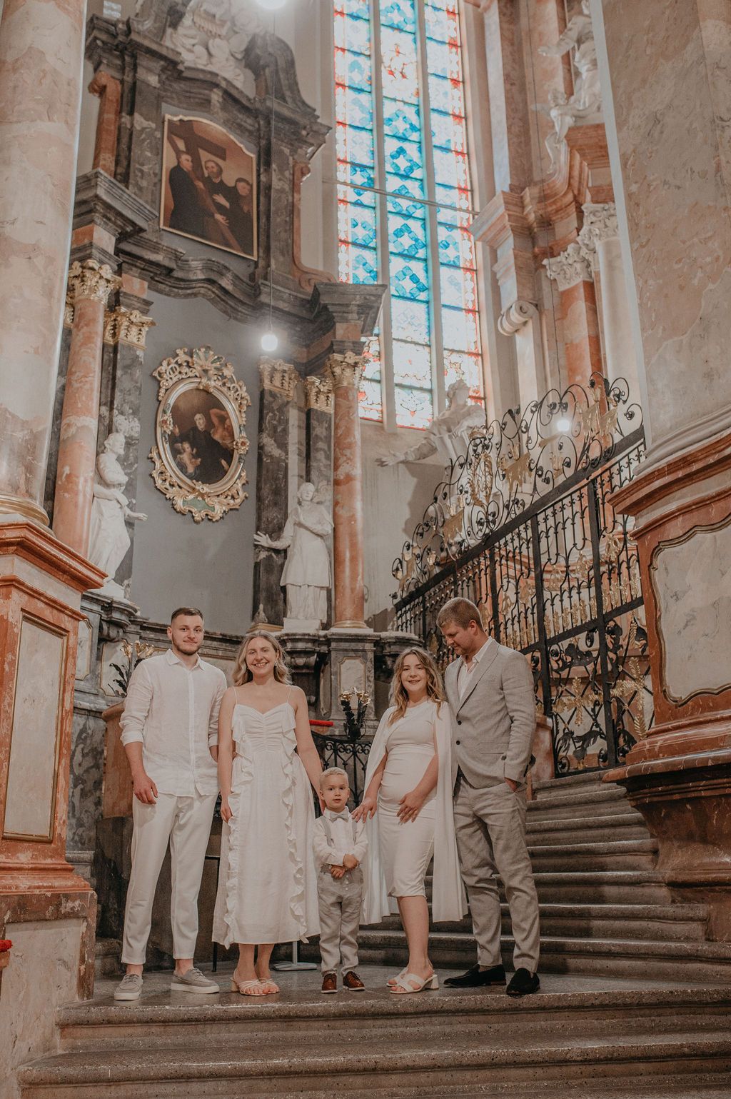 Family in white attire posing on church steps with ornate baroque altar and stained glass windows.