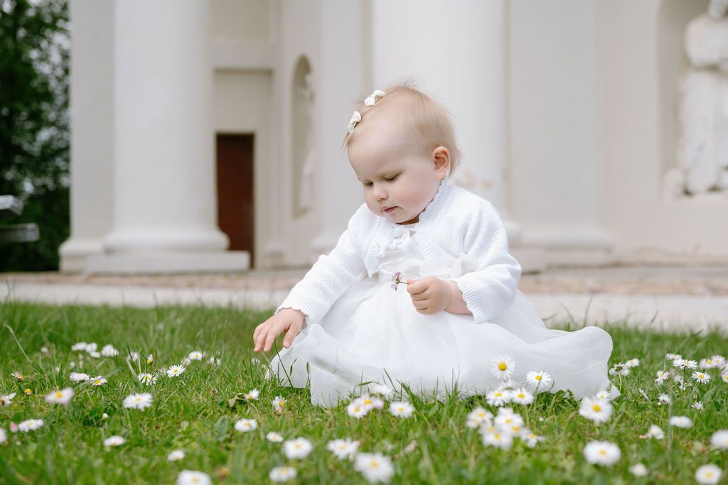 Baby in white dress sitting on grass with daisies near white columns.