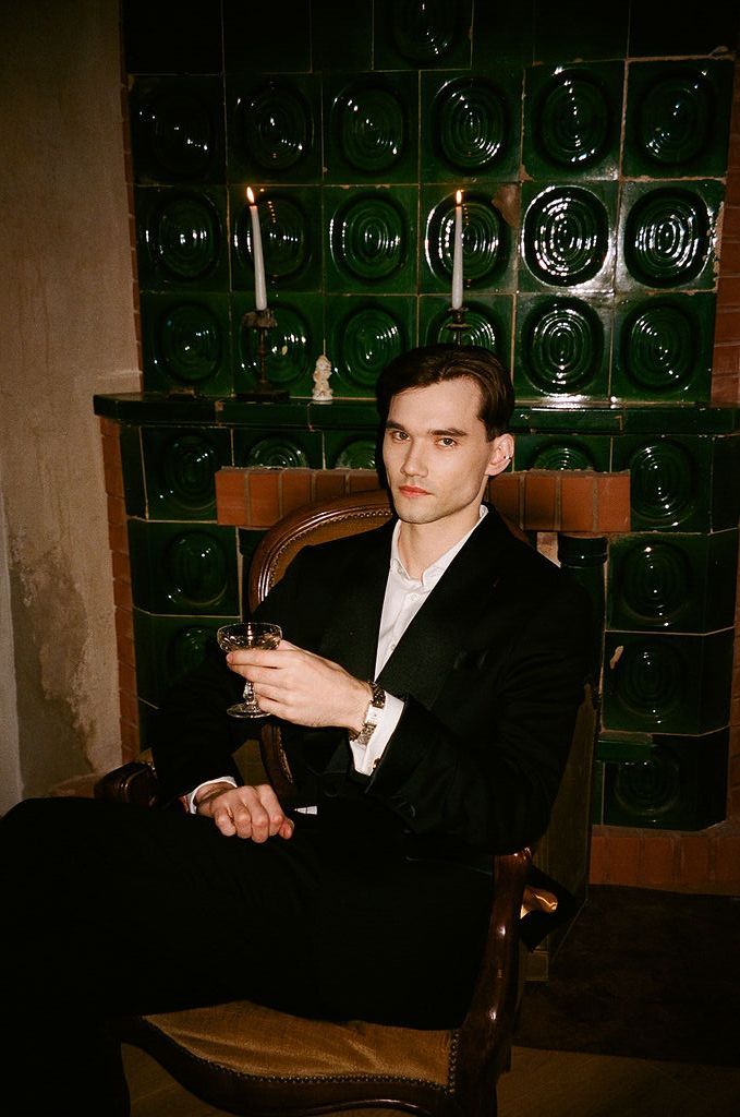 Young man in black suit holding a cocktail glass, seated in wooden chair against green tiled wall with candles.