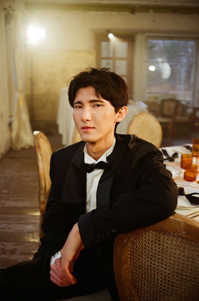 Young man in black tuxedo with bow tie seated at a dining table in a dimly lit elegant room.