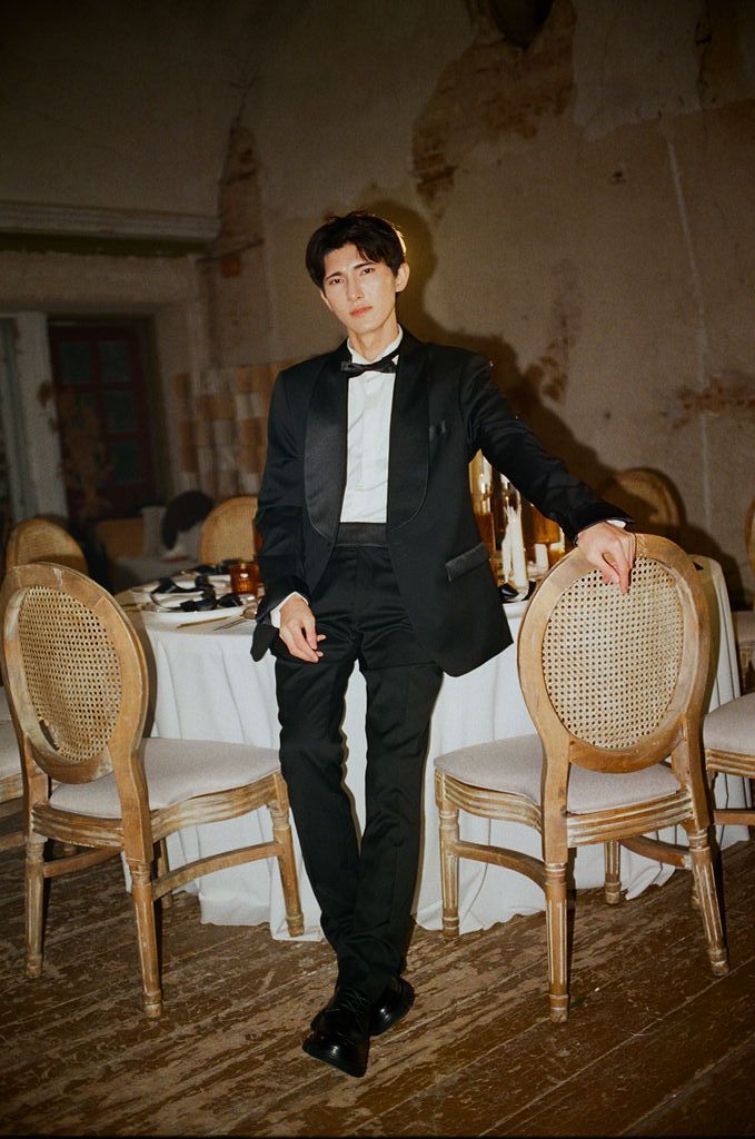 Stylish man in black tuxedo posing beside rattan chairs at a formal dining event.