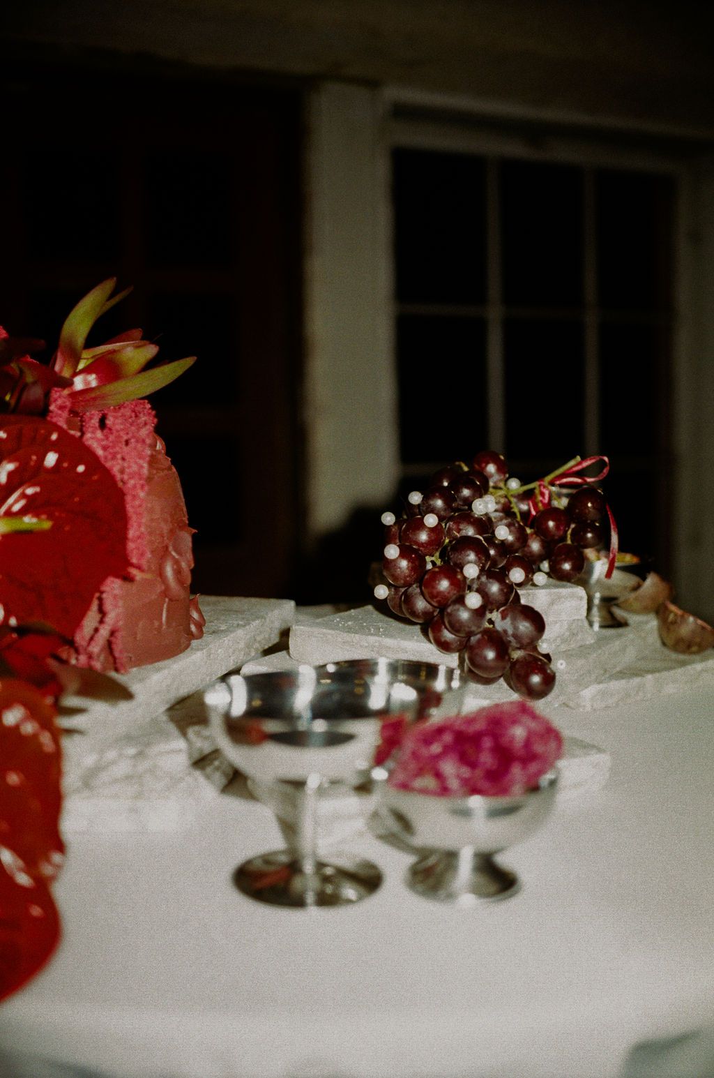 Silver chalices and dark red grapes displayed on a white lace tablecloth beside a red floral arrangement.