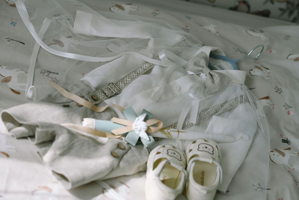 White baby baptism outfit with shoes, ribbons, and accessories laid flat on a patterned fabric surface.