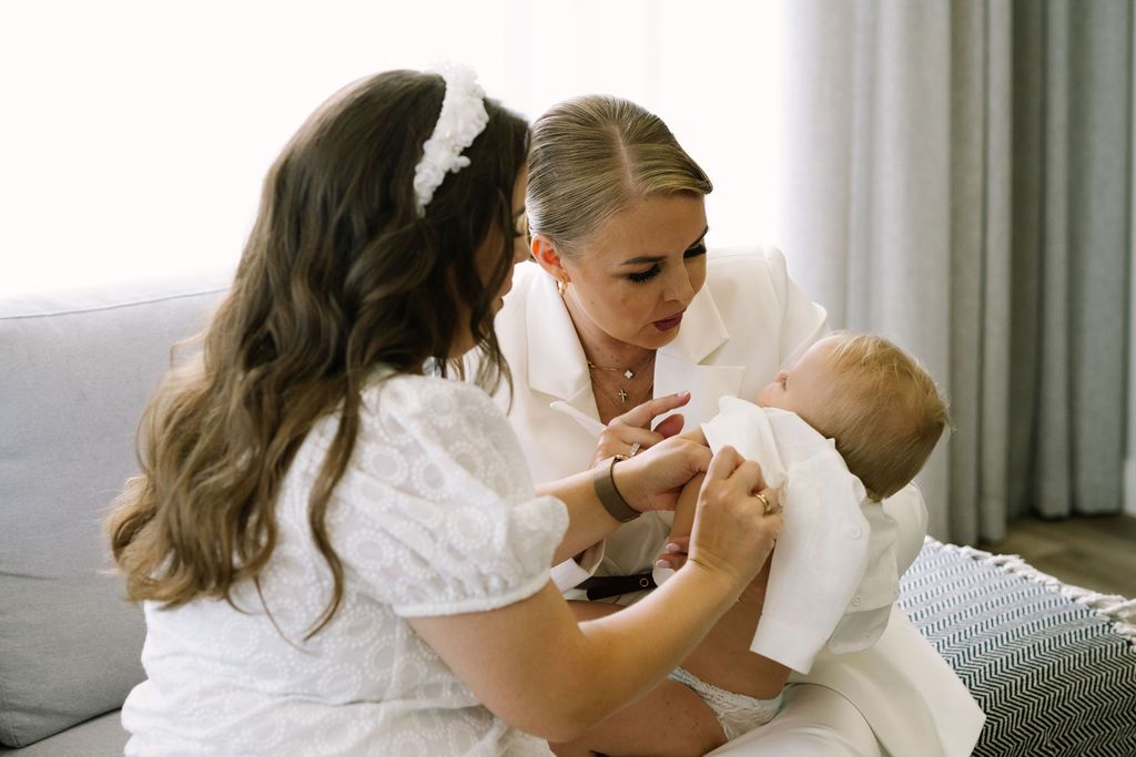 Two women in white dresses tenderly interact with a baby in white outfit on a cozy sofa.