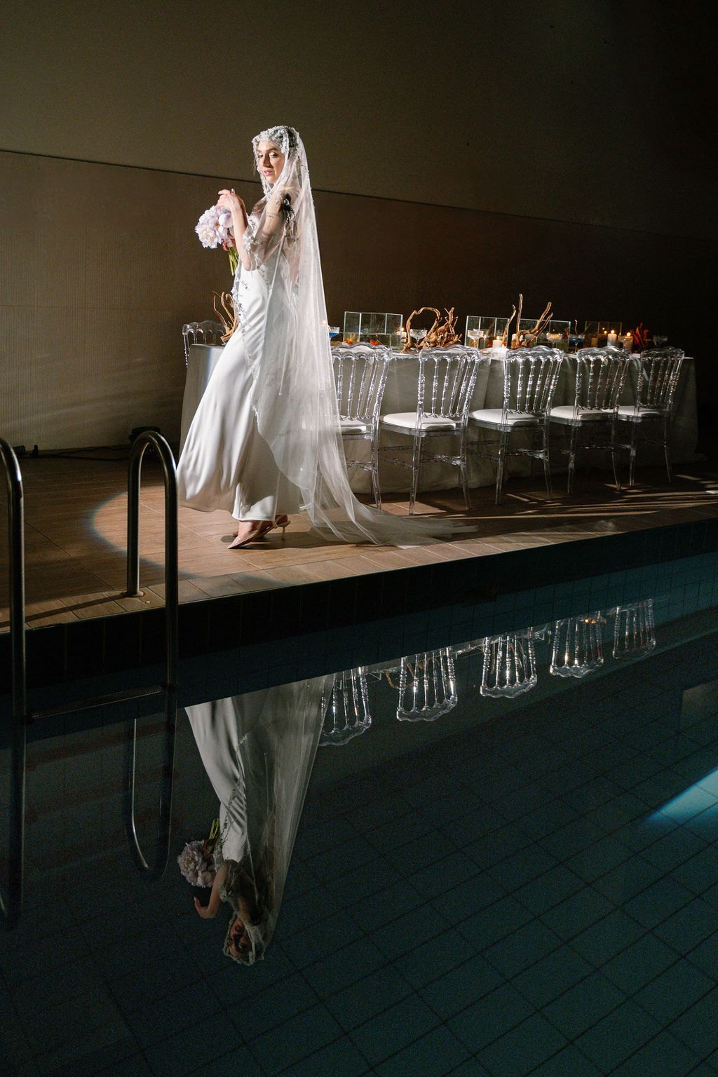 Bride in white gown and veil stands poolside near elegant reception table with clear chairs, her reflection mirrored in still water.