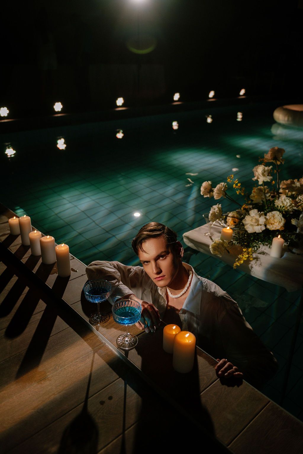 Aerial view of young man at candlelit poolside table with blue drinks and flowers at night.