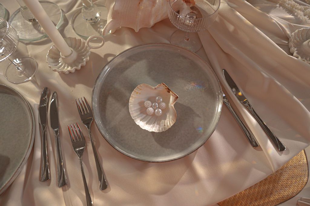Elegant table setting with a seashell on a silver charger plate, surrounded by silverware and soft pink linens.