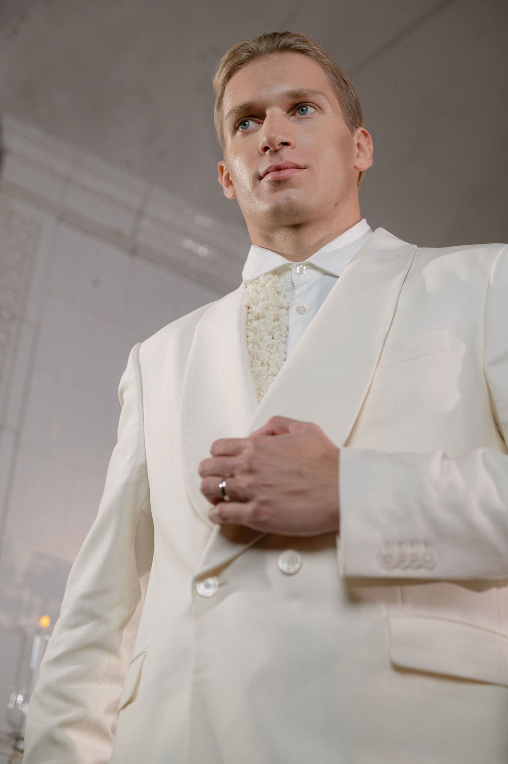 Elegant groom in ivory double-breasted suit with floral tie, posing confidently at a wedding venue.