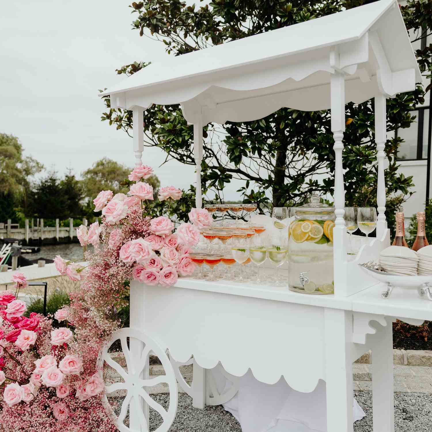 A white wedding dessert cart decorated with pink flowers and scalloped details stands outdoors in a garden setting.