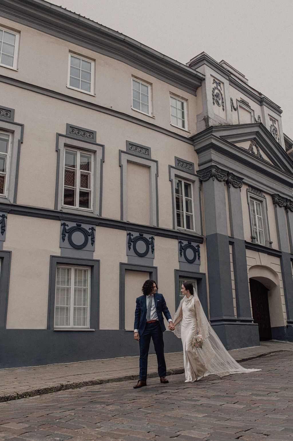 Bride and groom holding hands in front of a grand European-style building on cobblestone streets.