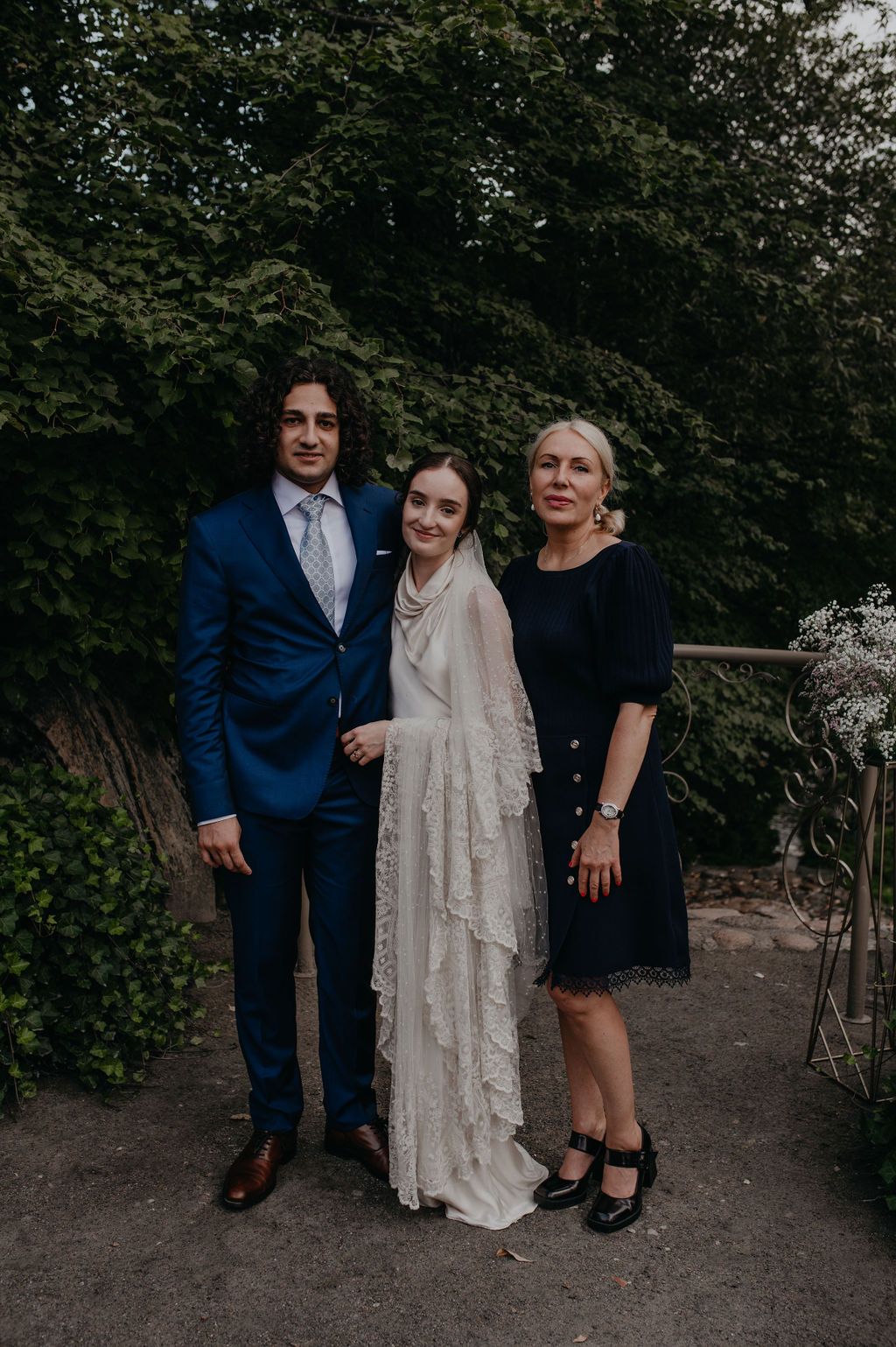 A bride in a white lace gown poses with a man in a blue suit and a woman in a navy dress outdoors.