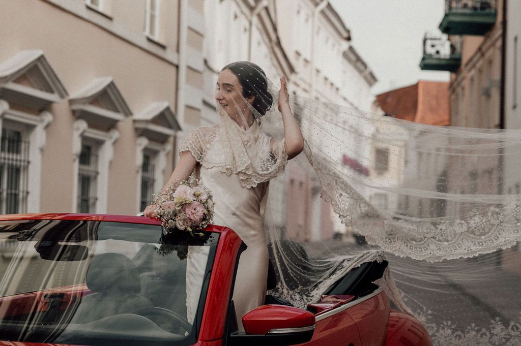 Bride in white dress standing in red convertible car, veil blowing in wind, holding bouquet on cobblestone street.