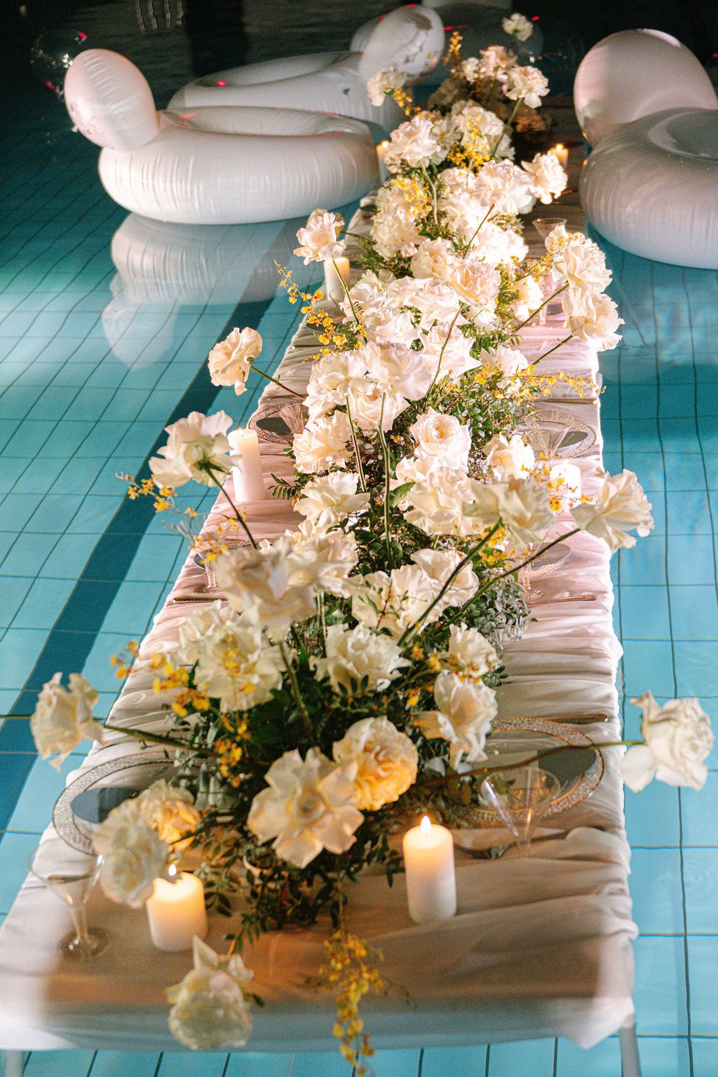 White and cream flowers with lit candles on a draped table set over a turquoise pool, with white inflatable floats nearby.