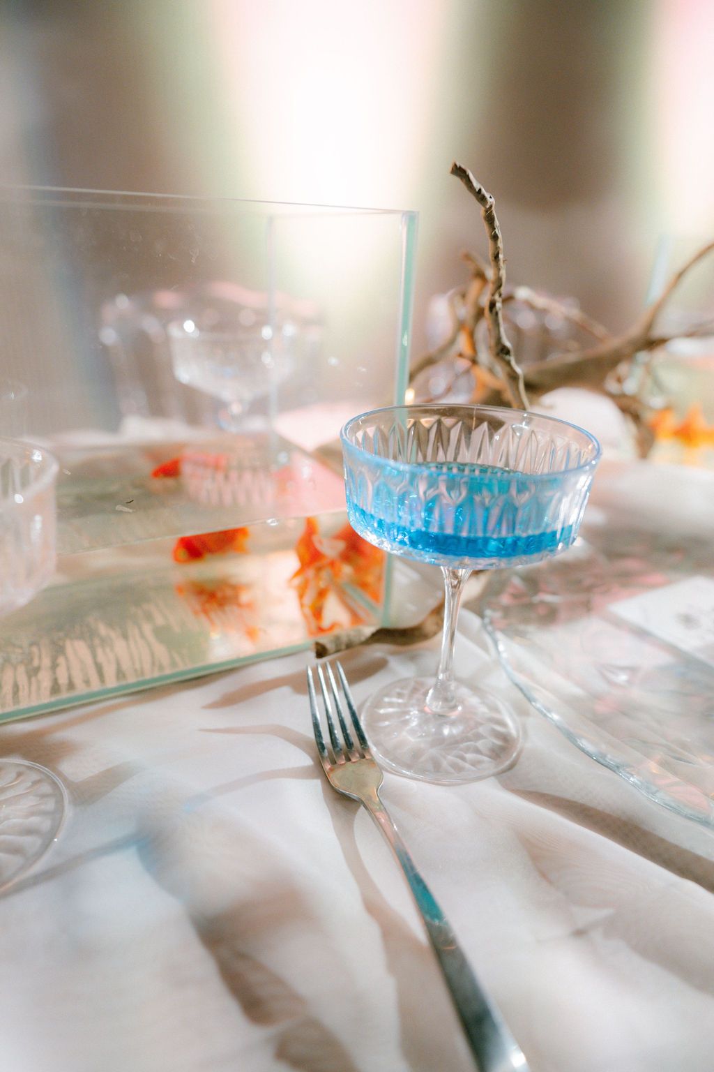 Blue cocktail in a crystal coupe glass on a white tablecloth beside a fork, a glass tank with goldfish and a dried branch.