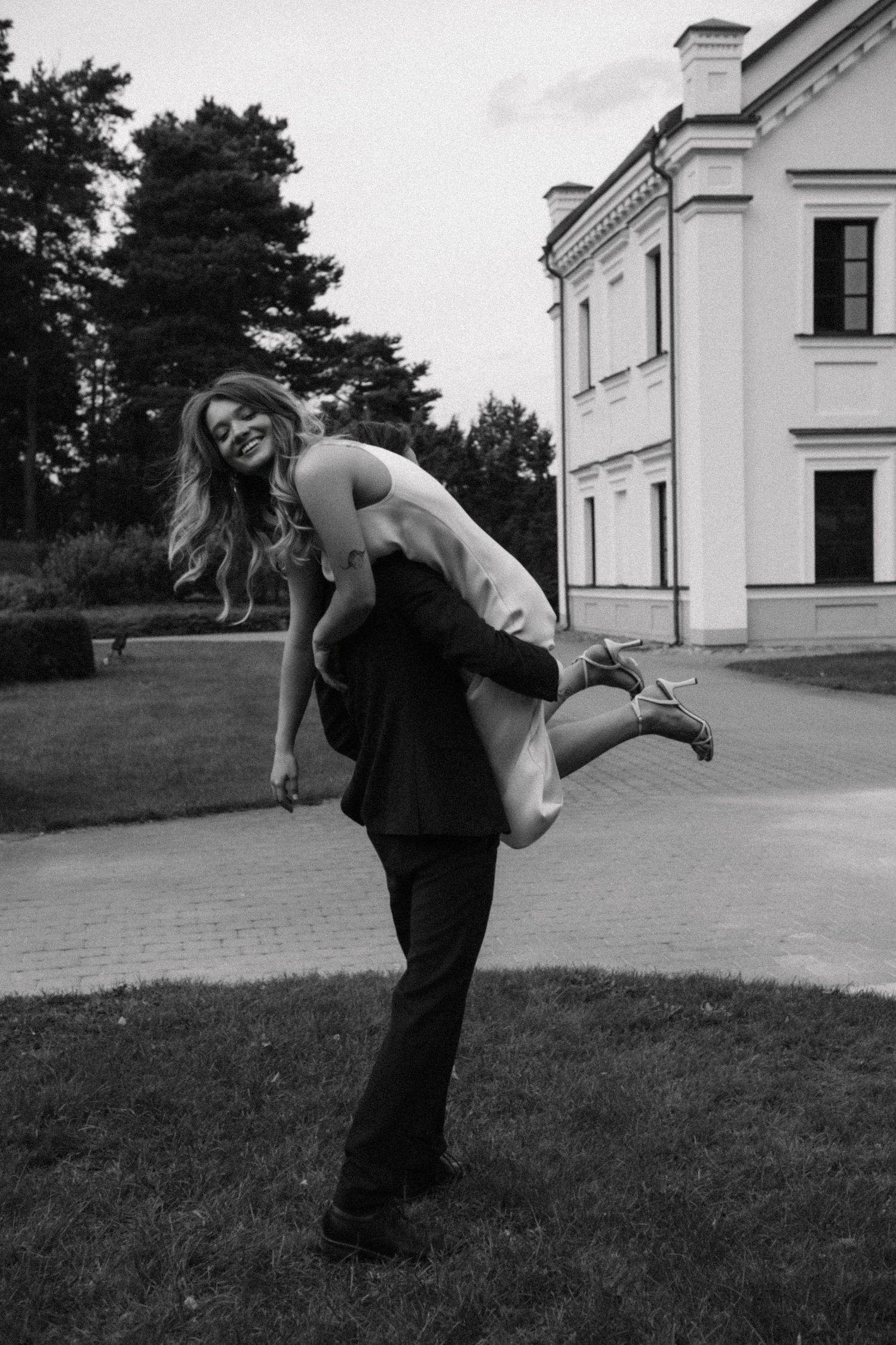 Black and white photo of a smiling woman in heels lifted playfully over a man's shoulder on a lawn in front of a classic building.