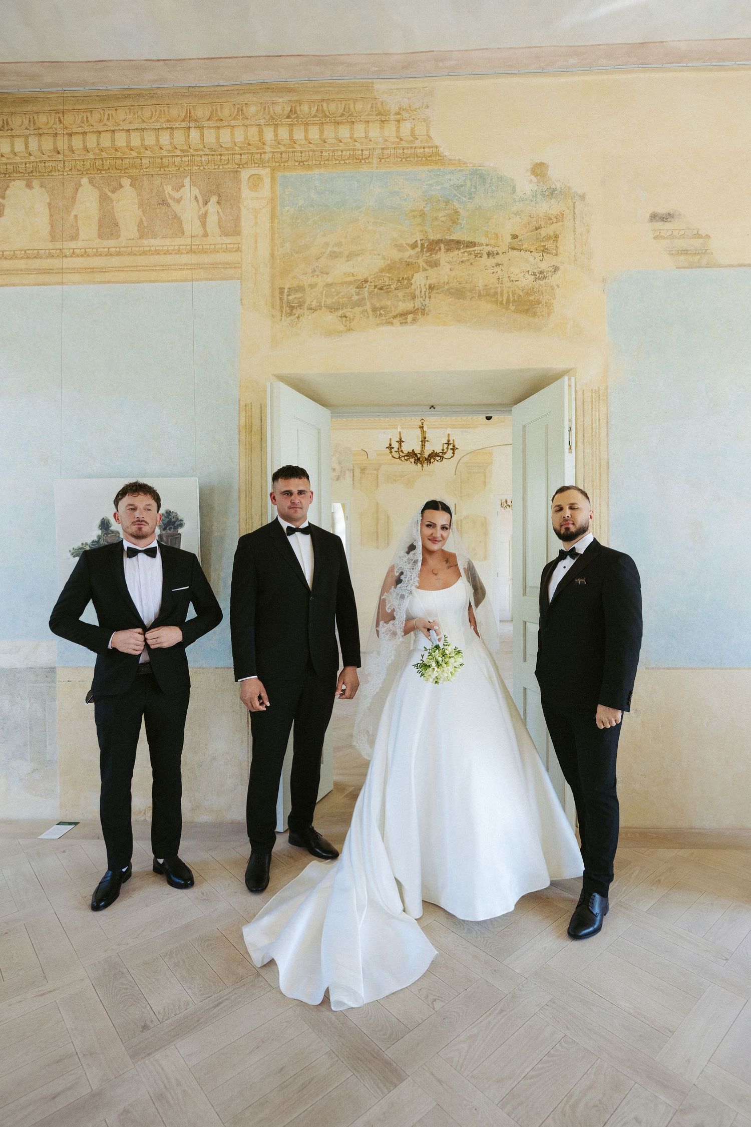 Bride in a white gown and veil holding a bouquet, posing with three groomsmen in black tuxedos inside a frescoed historic hall.