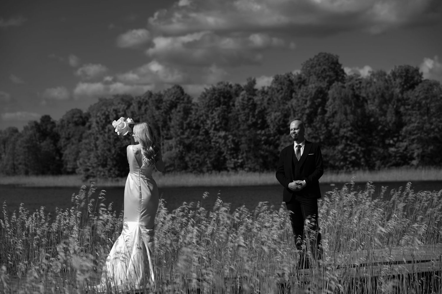 Black and white photo of a bride in a lace gown and a groom in a suit standing apart in tall grass beside a lake with trees behind.