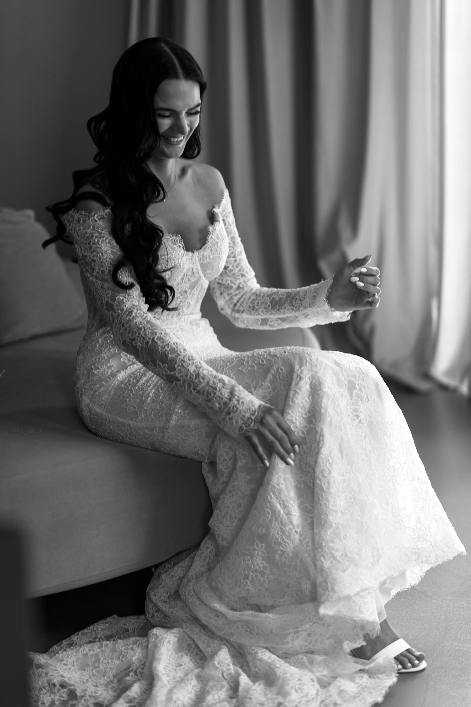 Black and white photo of a smiling bride in a lace long-sleeve wedding gown sitting elegantly on a sofa.