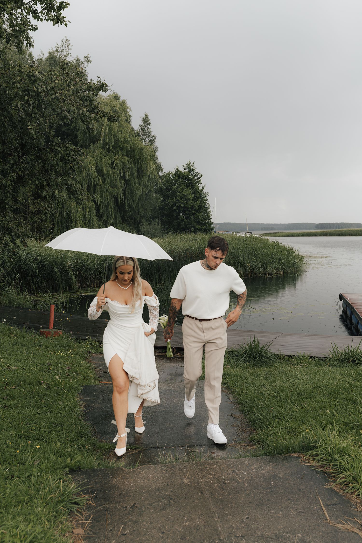 A couple walks along a lakeside path on an overcast day, with the person in white holding a white umbrella.
