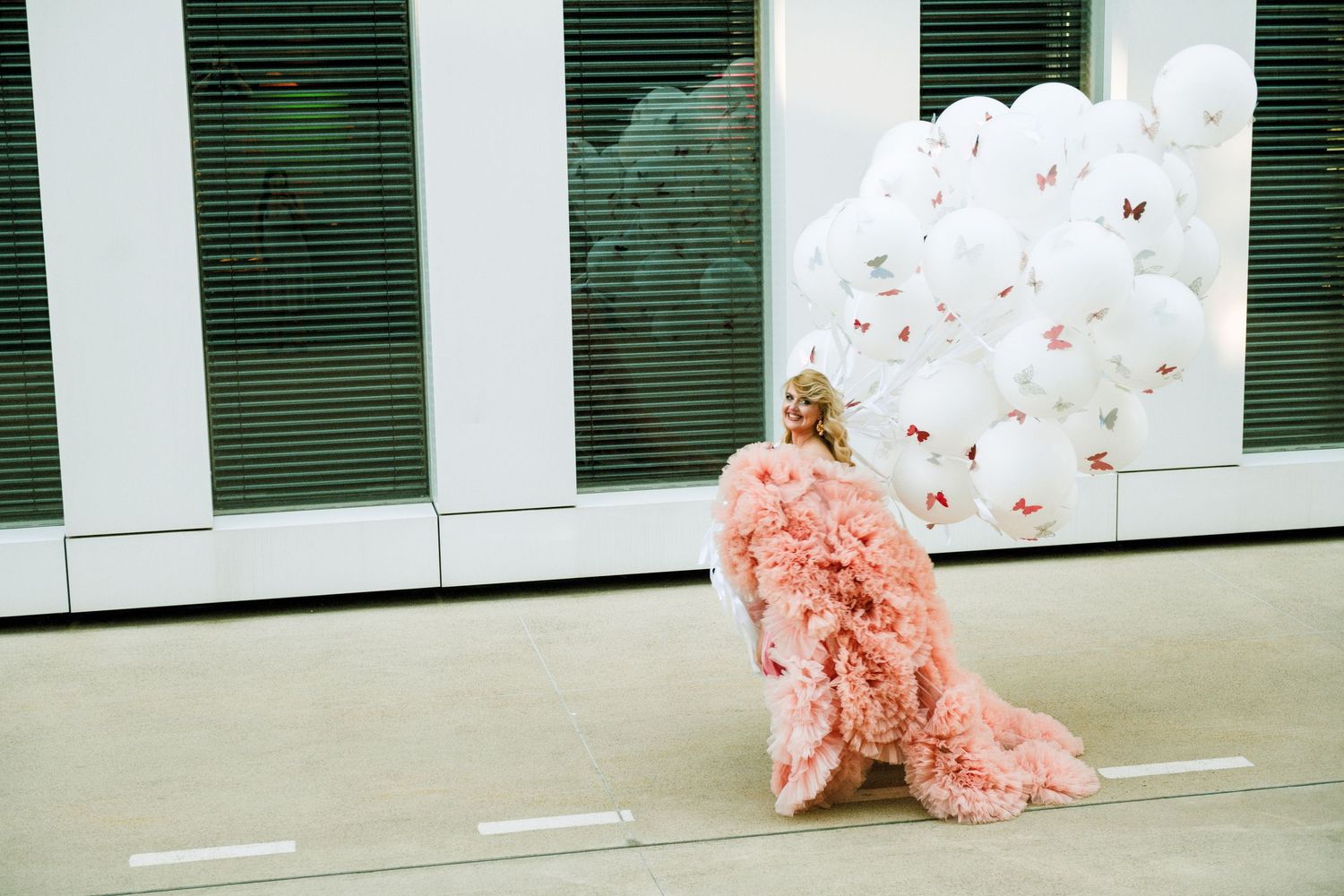 Woman in pink ruffled gown holding white butterfly balloons outdoors near a modern building.