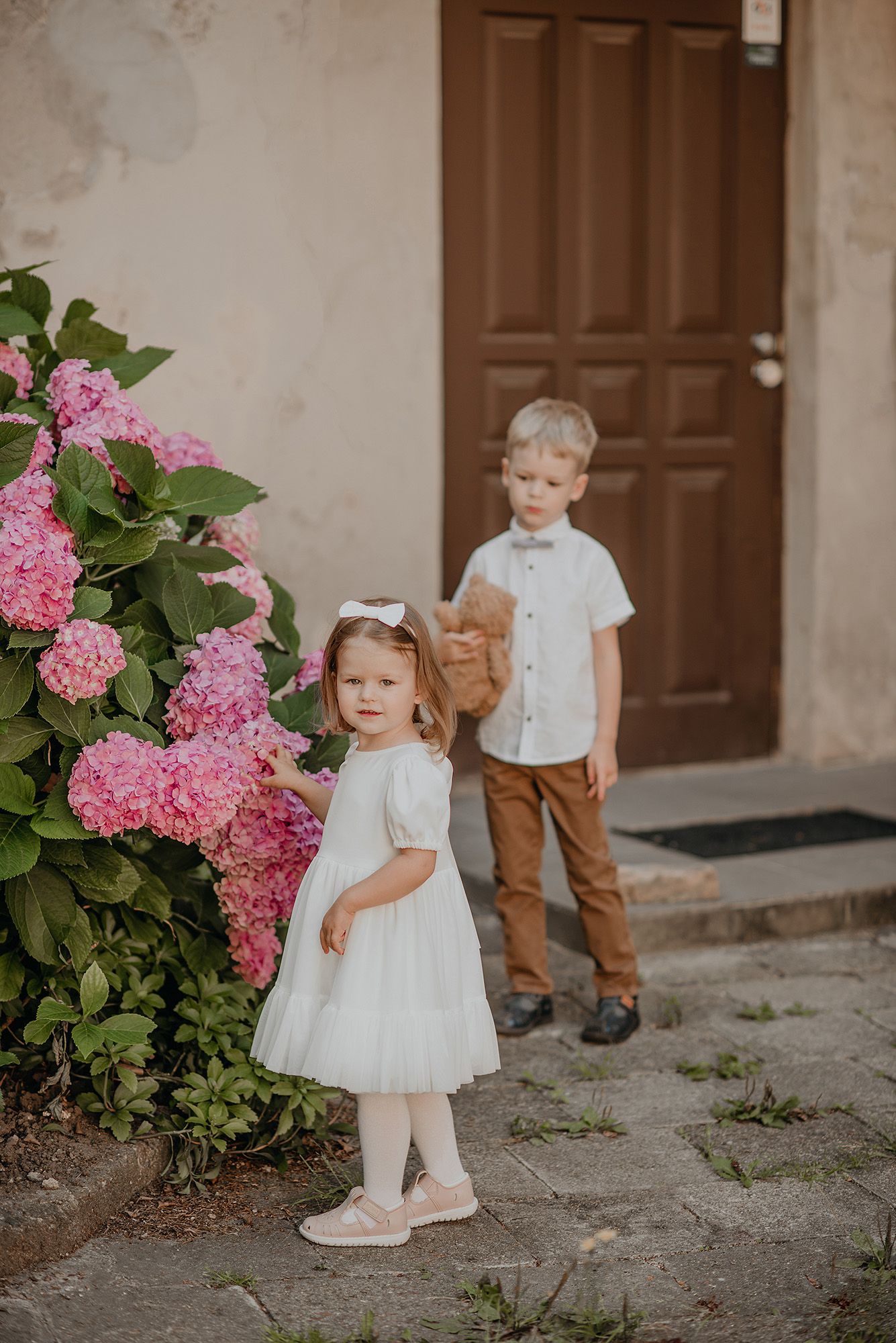 Two young children in coordinating outfits stand near pink hydrangea flowers on a stone patio.