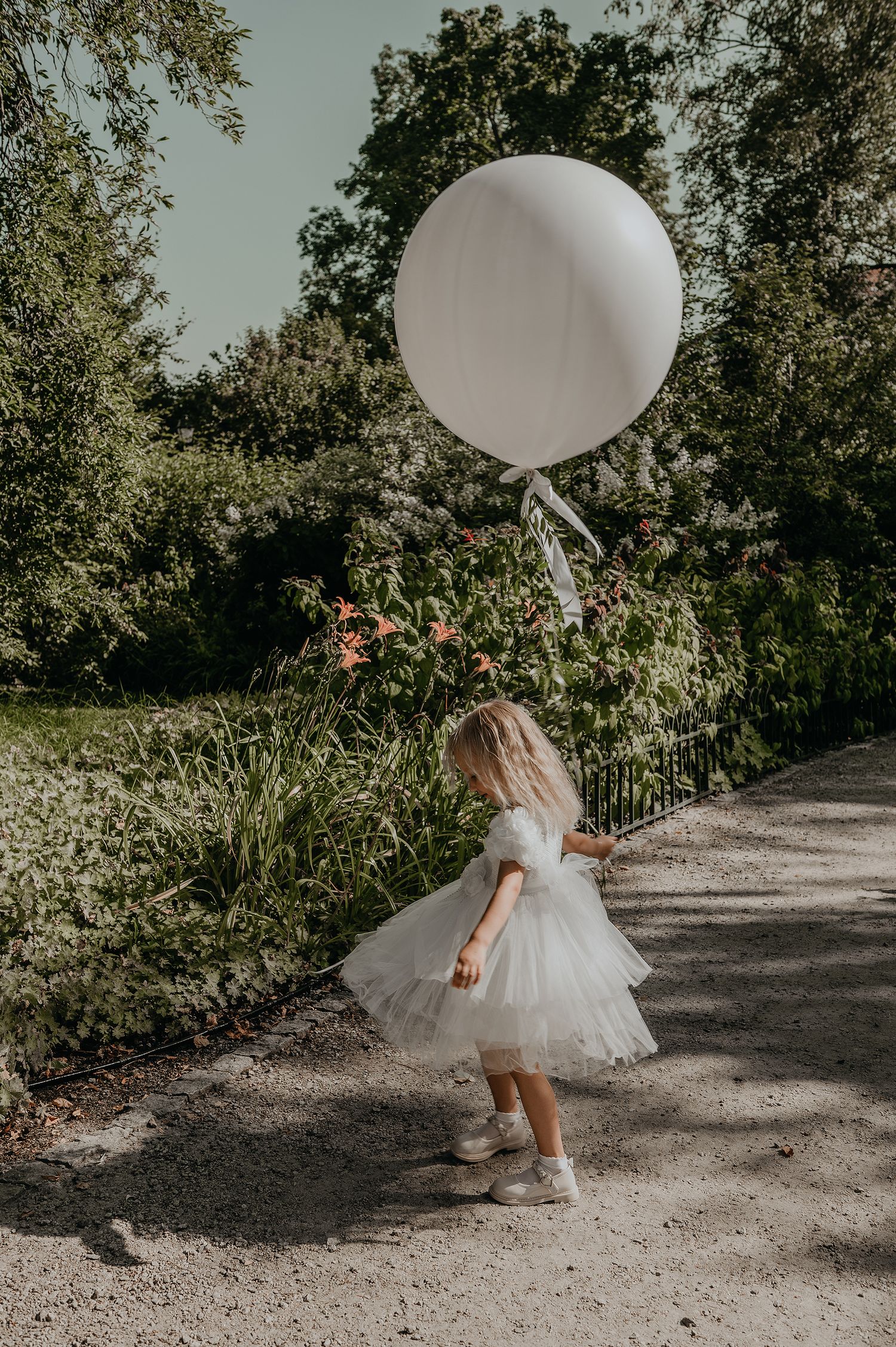 A dreamy garden scene with a white tulle dress and large white balloon on a winding path among lush greenery.