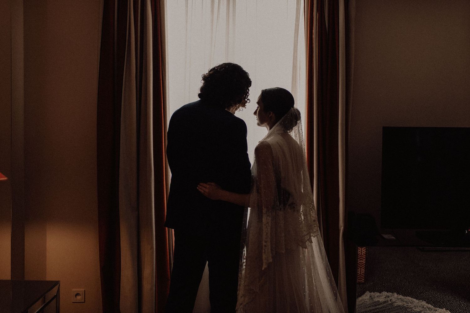 A couple in wedding attire stands in a dimly lit corridor with dramatic lighting creating a romantic atmosphere.