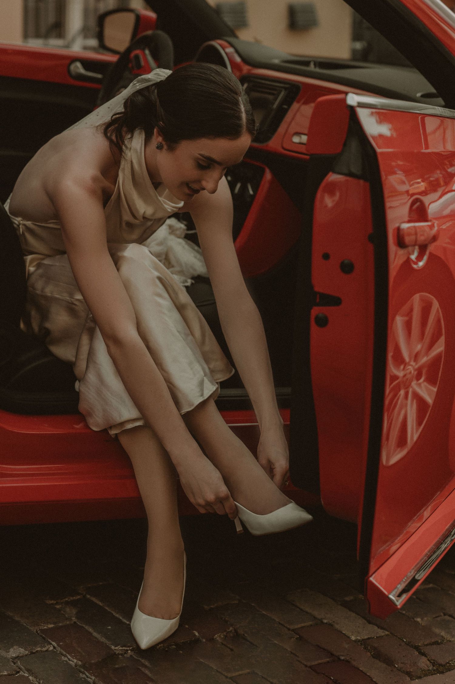 A bride in a white dress adjusts her shoes while sitting in a bright red vintage car.