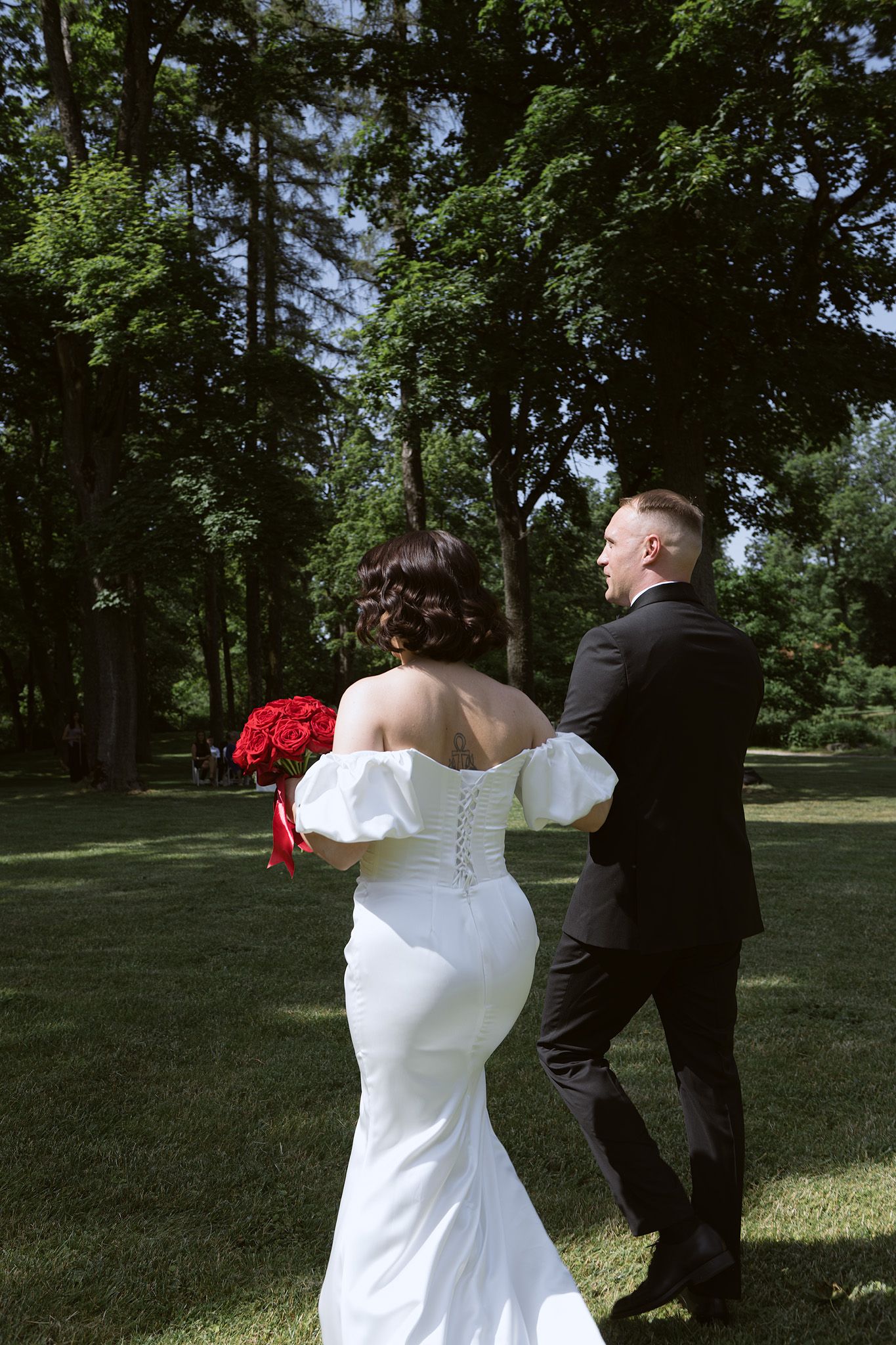 A couple in wedding attire walking together on a lush green lawn with red roses and trees in the background.