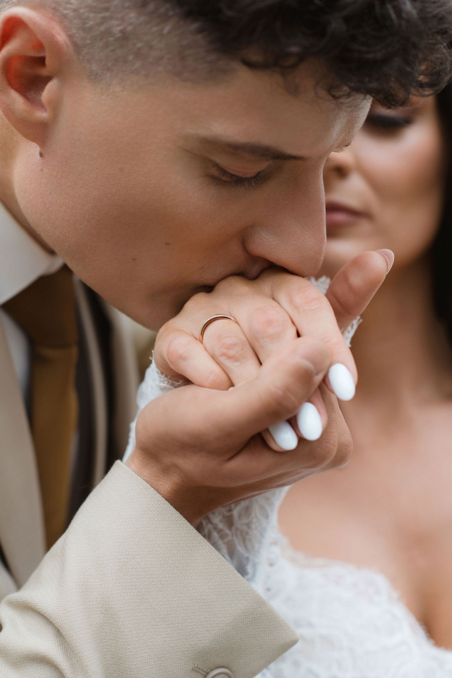 Groom in beige suit kissing bride's hand with wedding ring, romantic wedding moment close-up.