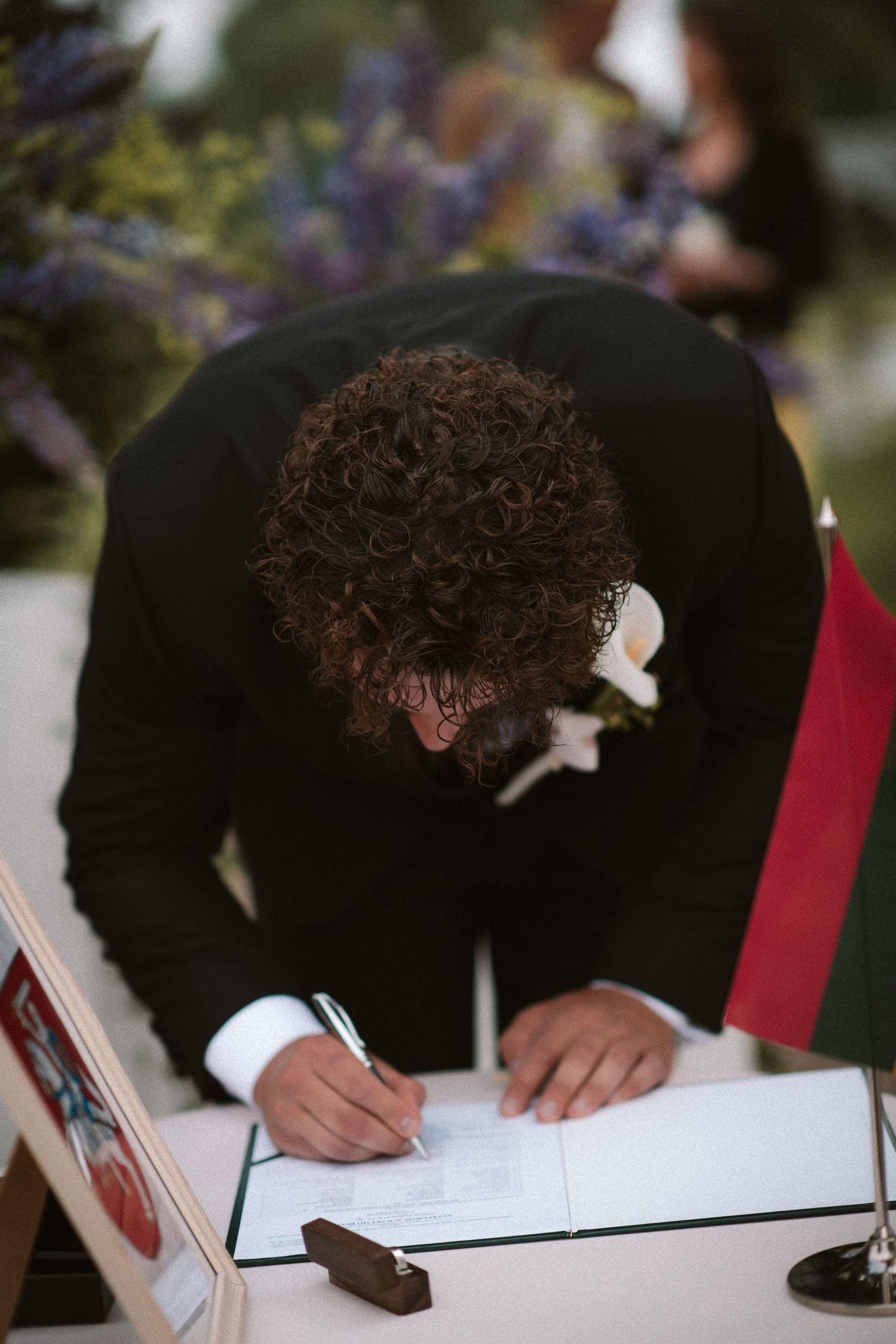 Groom in black suit signing wedding registry documents at outdoor ceremony table.