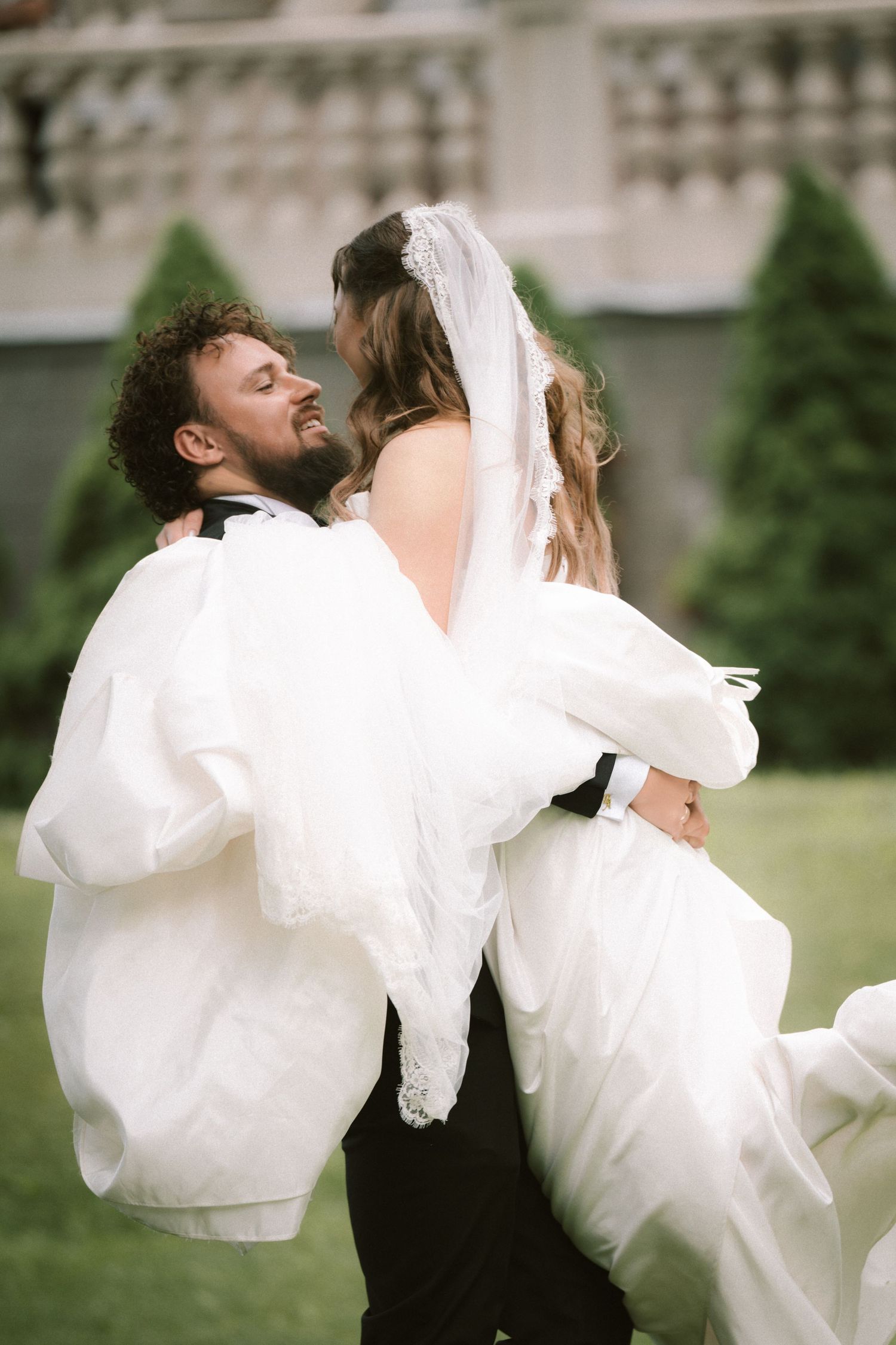 Joyful groom lifting bride in white wedding dress outdoors near elegant stone building.
