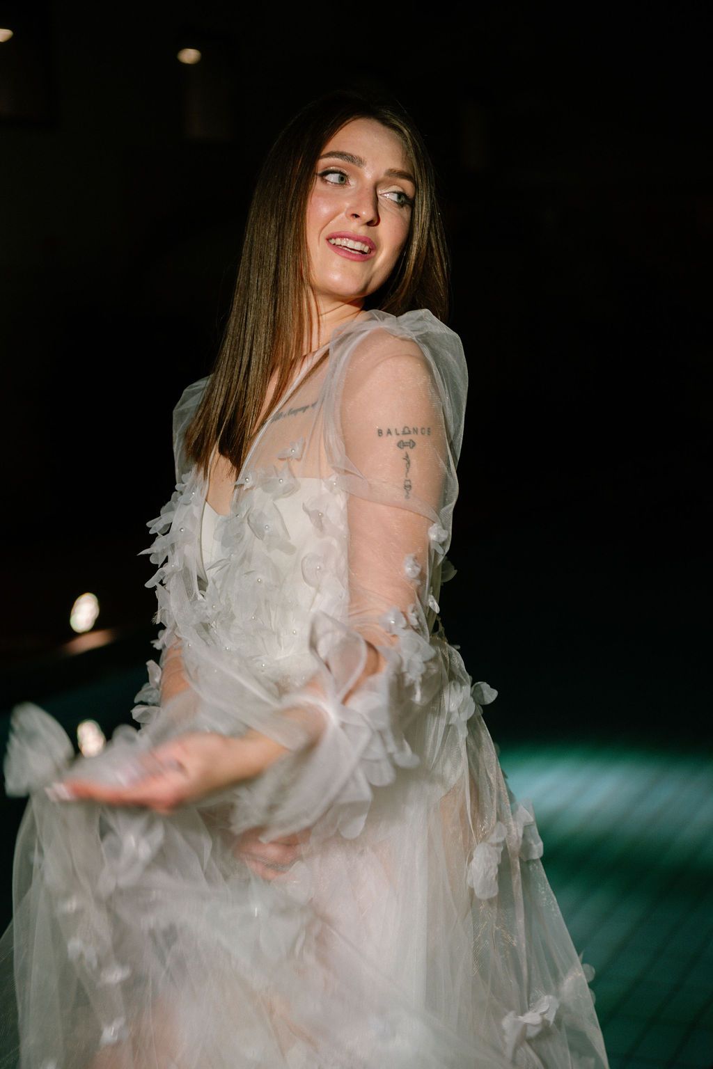 Woman with long brown hair smiles over her shoulder in a flowing white tulle gown with floral appliqués and sheer sleeves.