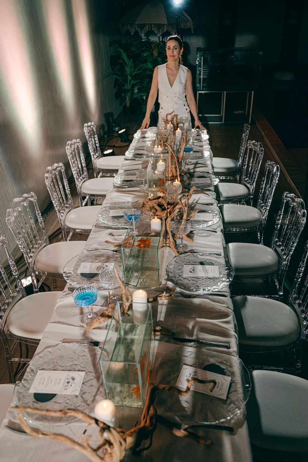 Bride in a white dress stands at the end of a long elegant wedding reception table with candles, driftwood centerpieces, and glass plates.