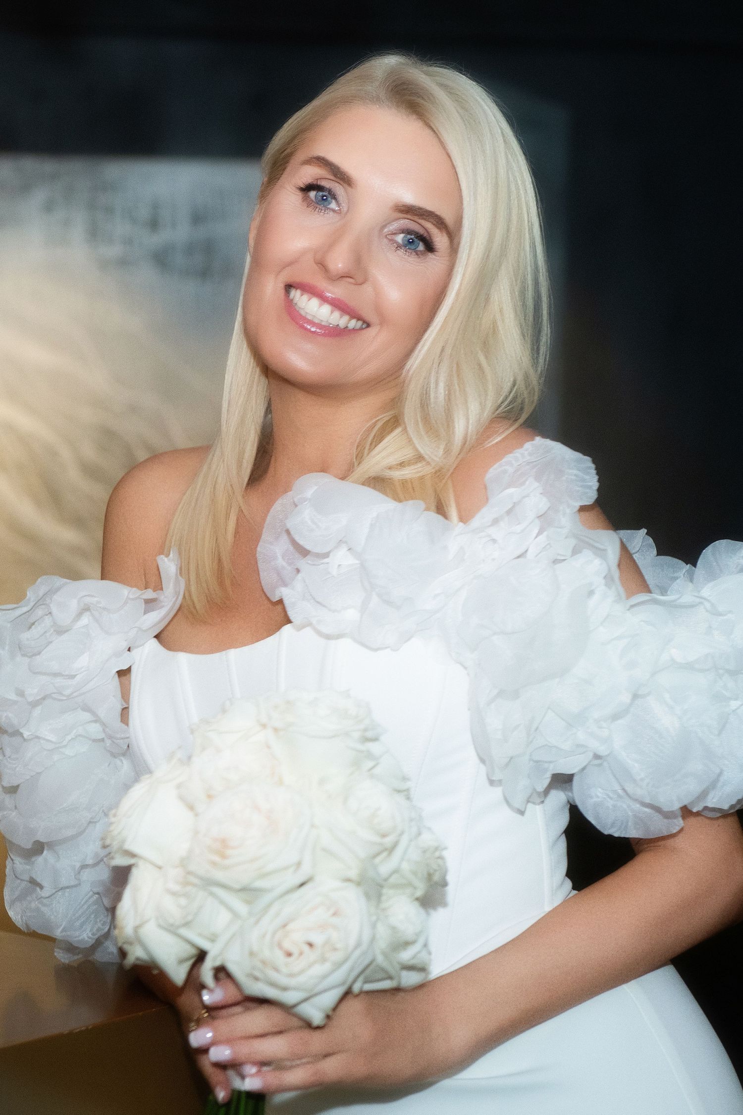 Smiling blonde bride in a white dress with ruffled off-shoulder sleeves holding a bouquet of white roses.