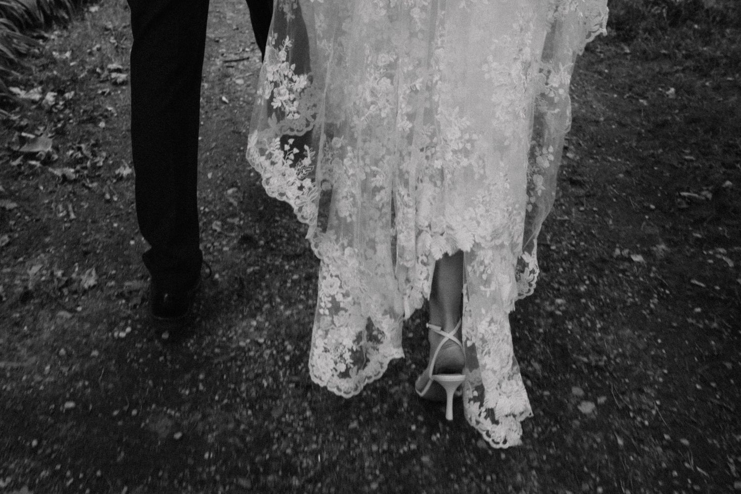 Black and white photo of a bride in a lace wedding gown and heeled sandals walking beside a groom in dark trousers on a dirt path.