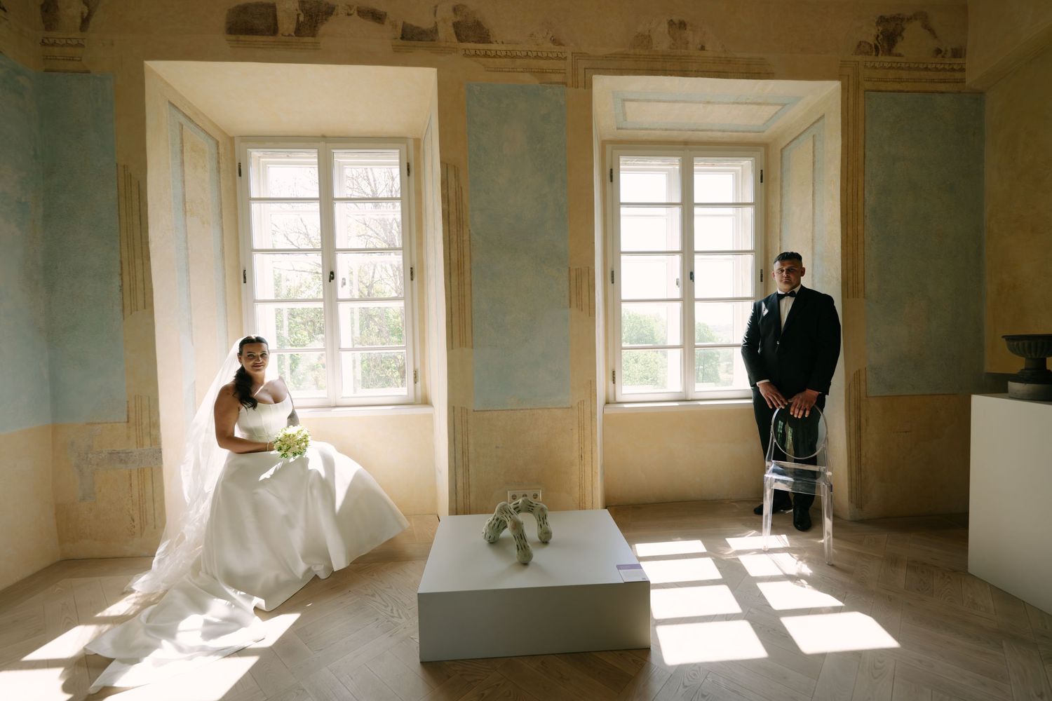 Bride in a white gown sits on a white bench while the groom in a dark suit stands by a chair in a sunlit room with tall windows.
