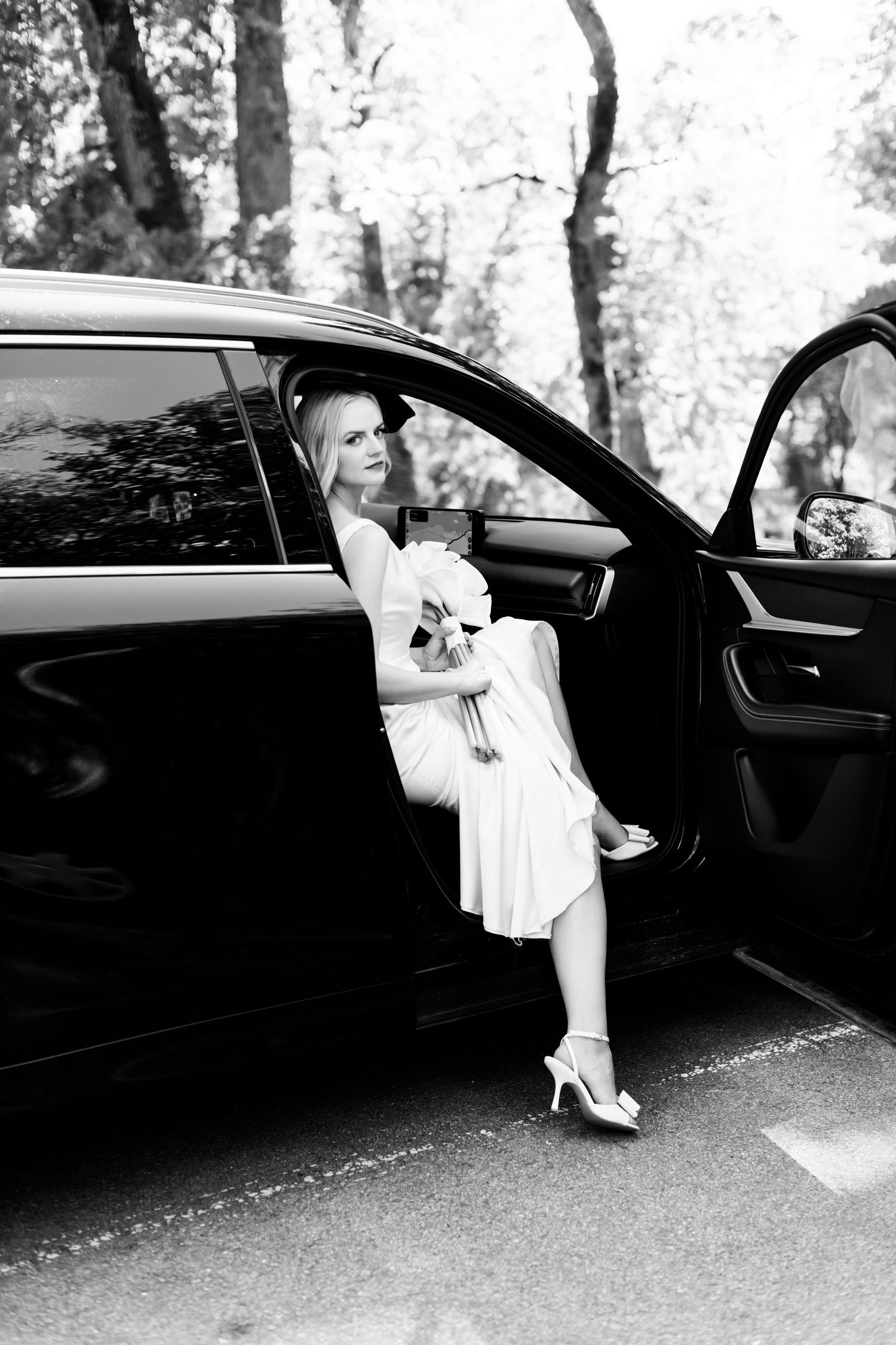 Black and white photo of a woman in a white dress and heels sitting in the open door of a black car, holding a bouquet.