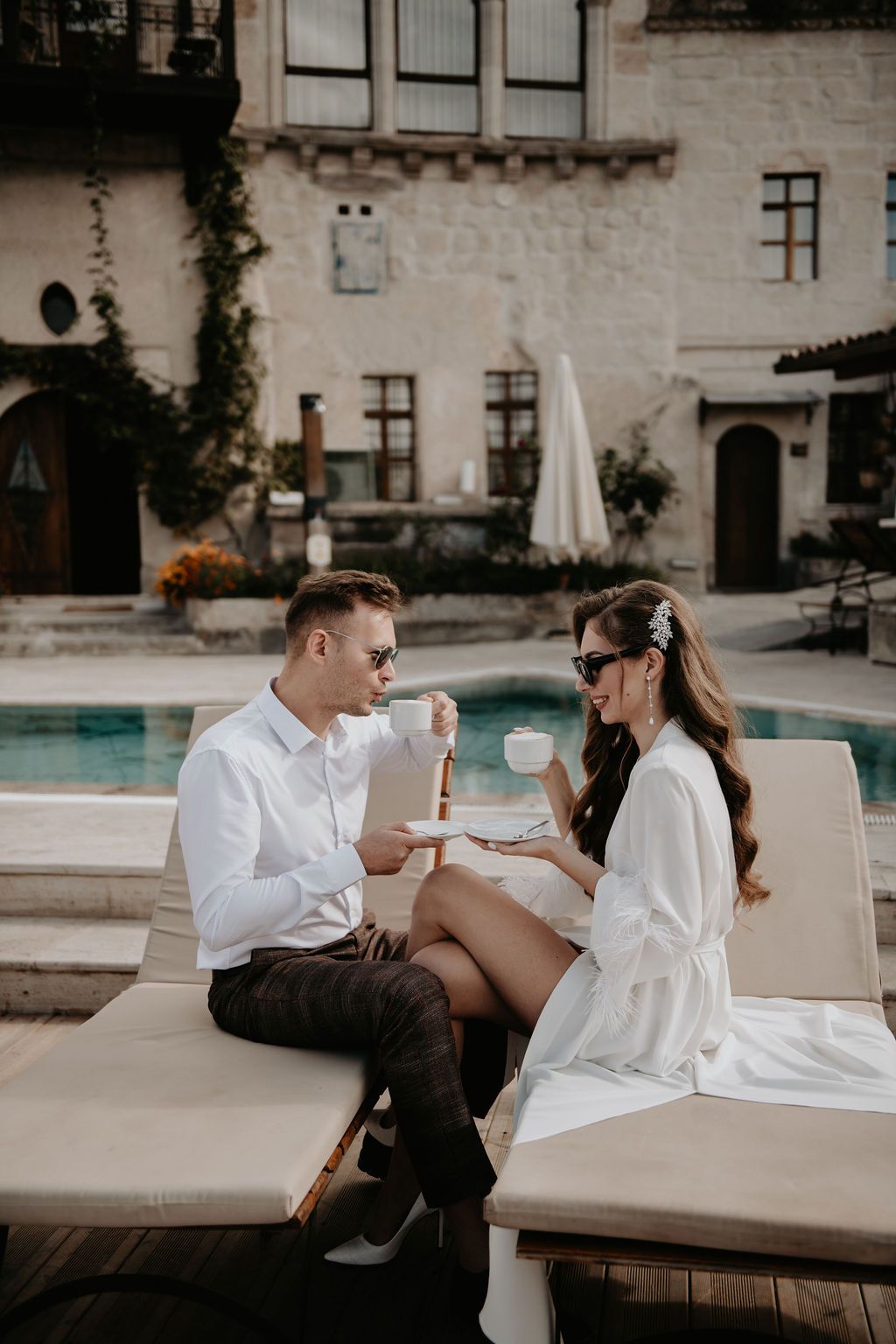 Elegant couple in white outfits enjoying coffee by a luxury pool at a stone villa resort.