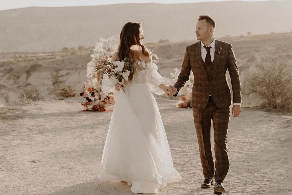 Bride and groom walking hand in hand through desert landscape with bridal bouquet.