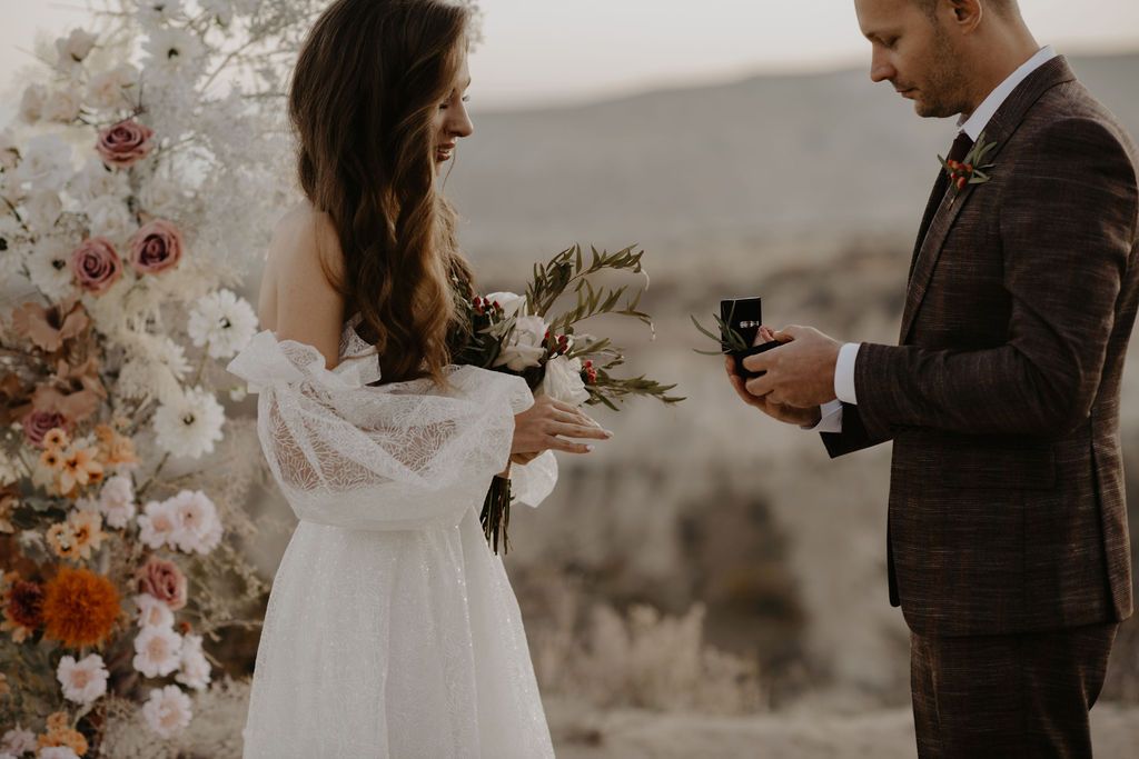 Bride in lace gown receiving ring from groom in brown suit before floral arch at outdoor ceremony.