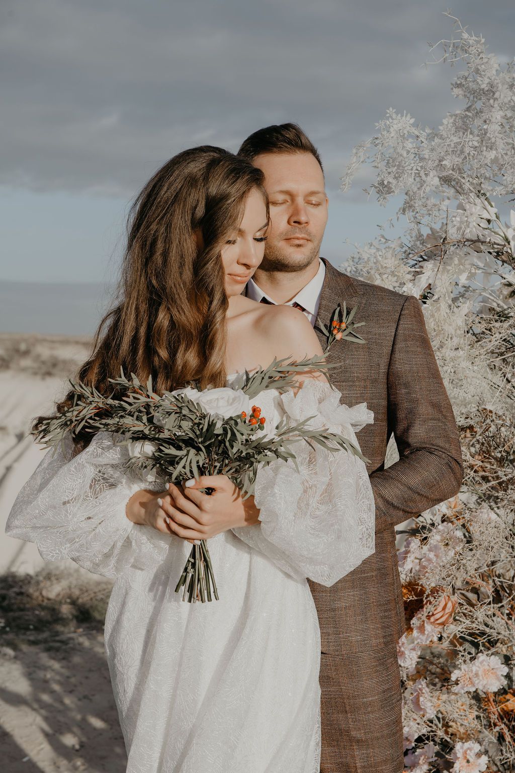 Bride in white dress holding bouquet embraced by groom in brown suit during outdoor wedding photoshoot.