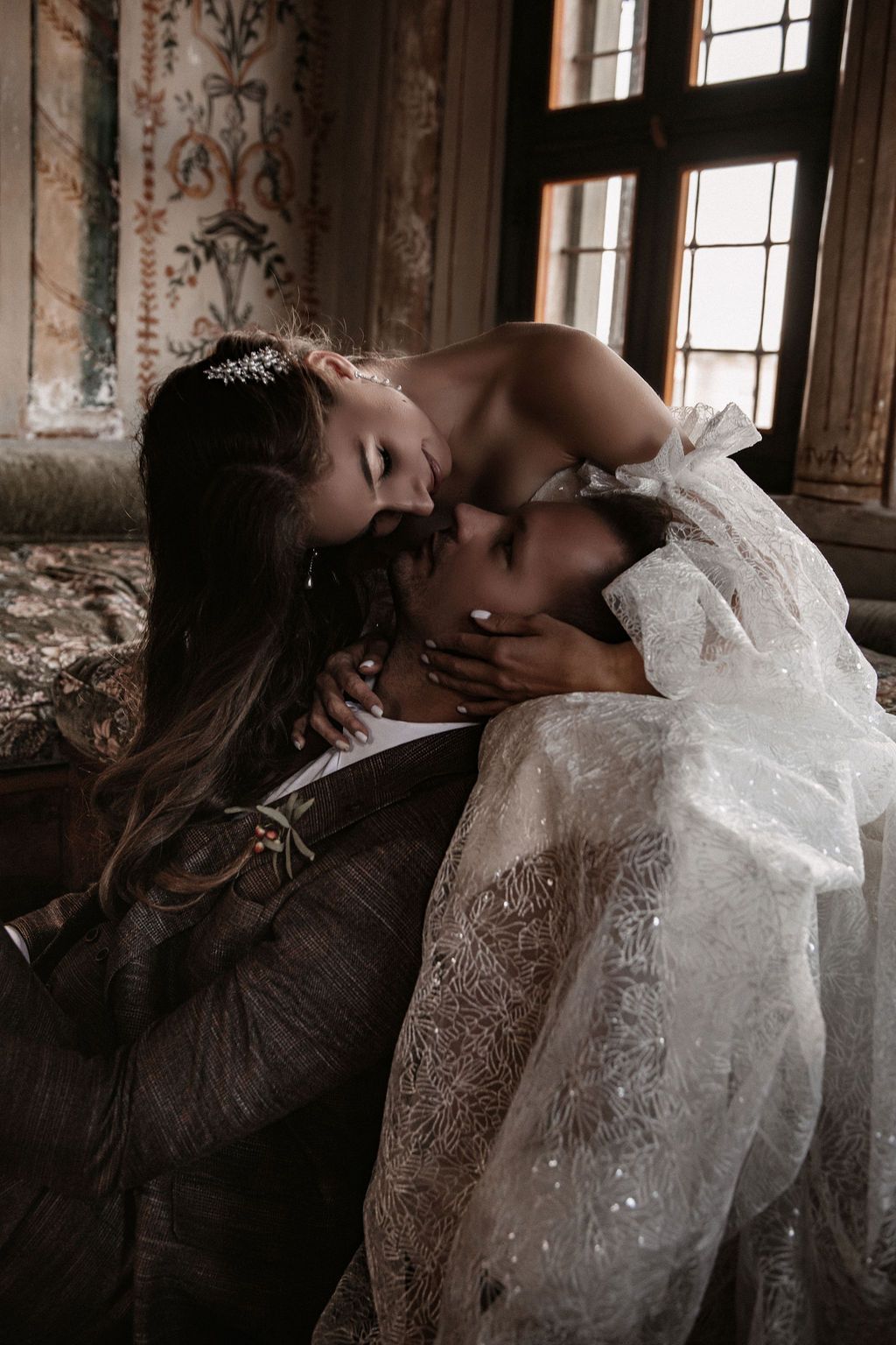 A bride in a lace gown and hair accessory leans in to kiss her groom on a vintage sofa in an ornate room.