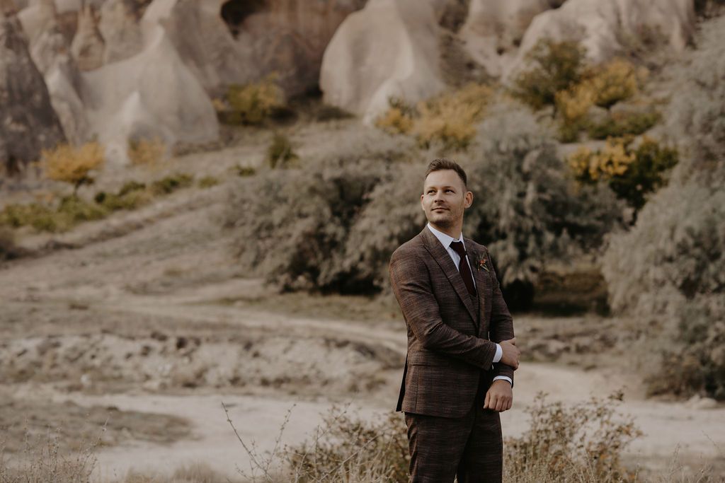 A well-dressed groom in a brown plaid suit stands confidently in a scenic Cappadocia landscape with rocky formations.