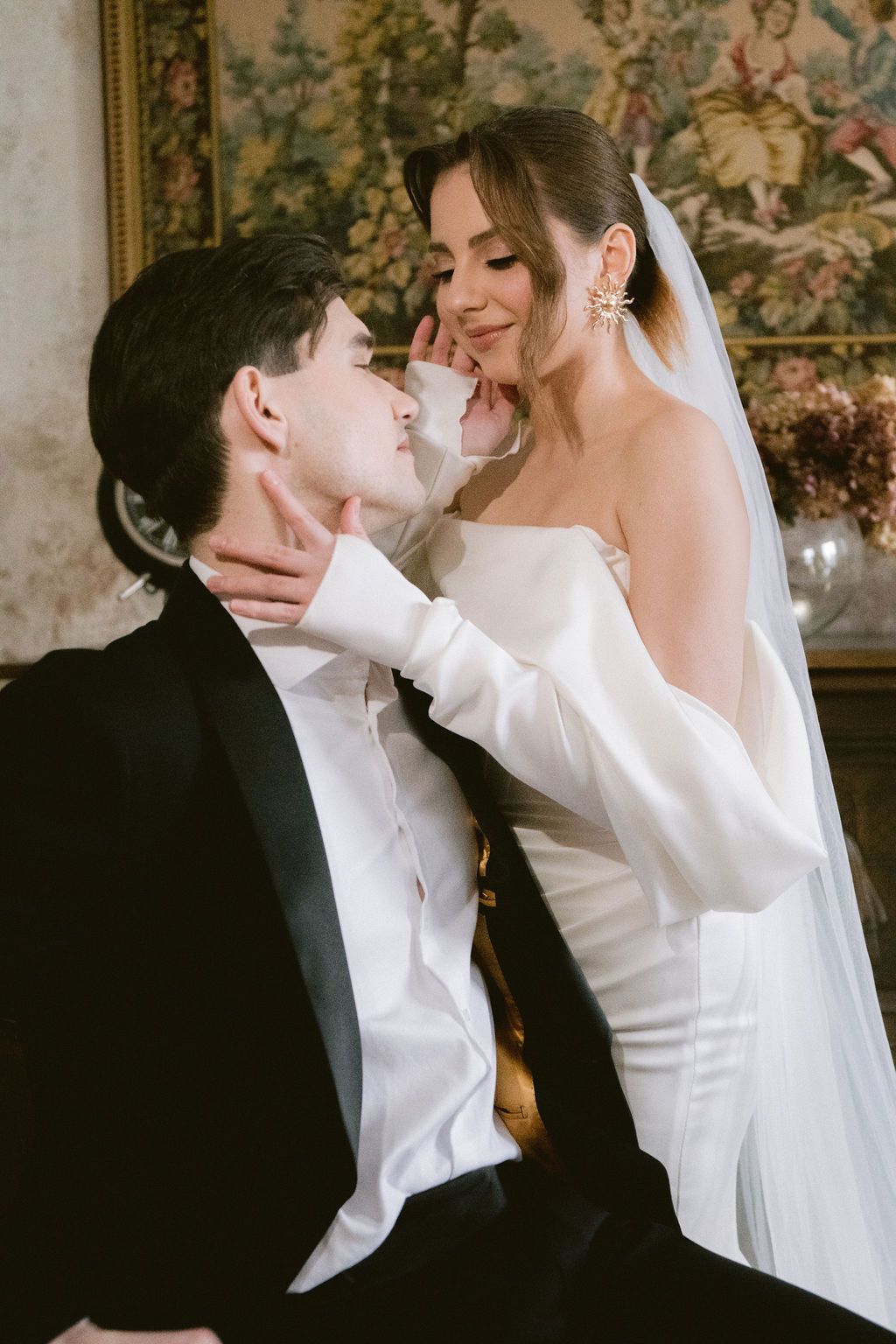 Bride in white off-shoulder gown and veil tenderly cups groom's face in black tuxedo during romantic wedding portrait.