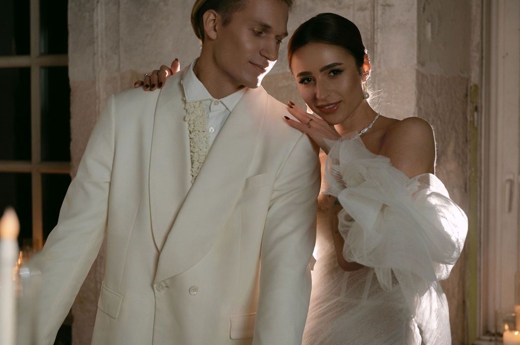 Elegant couple in white wedding attire, groom in suit and bride in off-shoulder gown, posing romantically indoors.